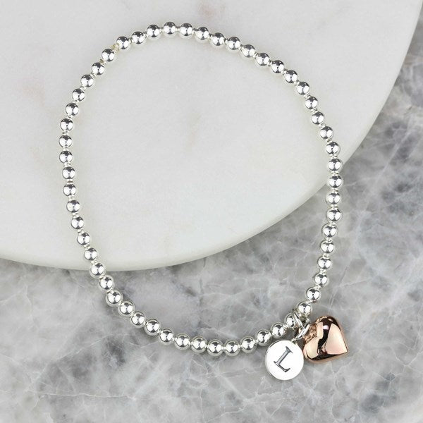 A silver beaded stretch bracelet with a small round silver disc charm engraved with the letter 'L' and a rose gold heart charm, displayed on a white ceramic plate over a gray marble surface.