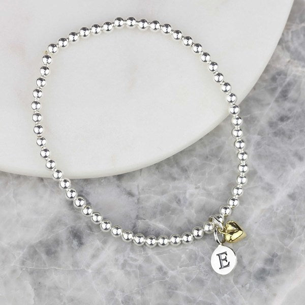 A silver beaded stretch bracelet with a small round silver disc charm engraved with the letter 'E' and a small gold heart charm, displayed on a white ceramic plate over a gray marble surface.