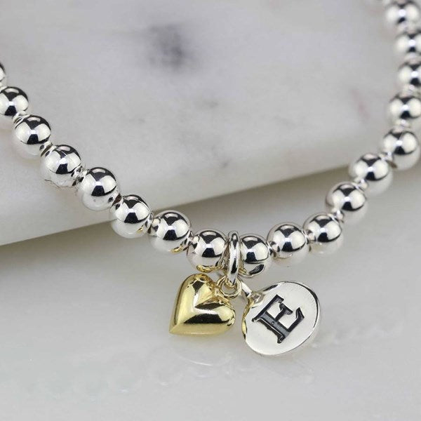 A silver beaded bracelet with two dangling charms: a polished gold heart and a silver oval disc engraved with a black capital letter H, displayed on a white marble surface.