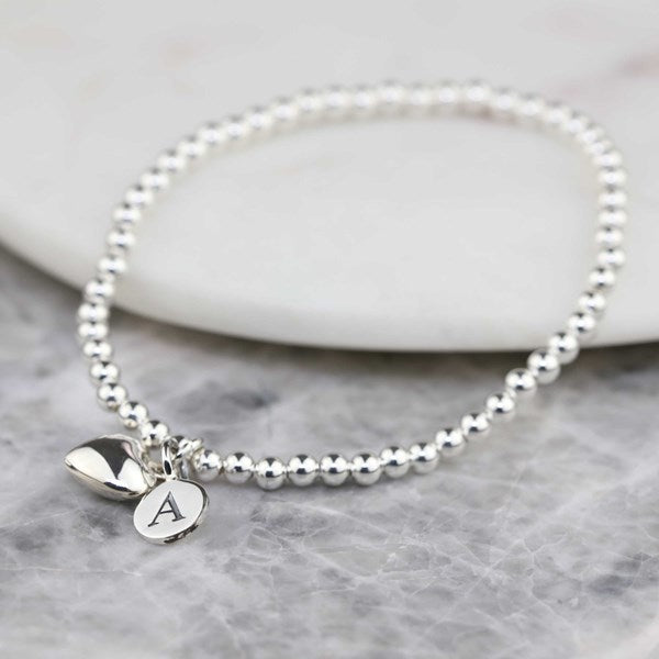 A silver beaded bracelet with two charms, one heart-shaped and one oval engraved with the letter 'A', displayed on a light marble surface.