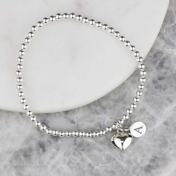 A silver beaded bracelet with two heart charms, one featuring a monogram letter 'A', displayed on a white marble slab over a gray marble surface.