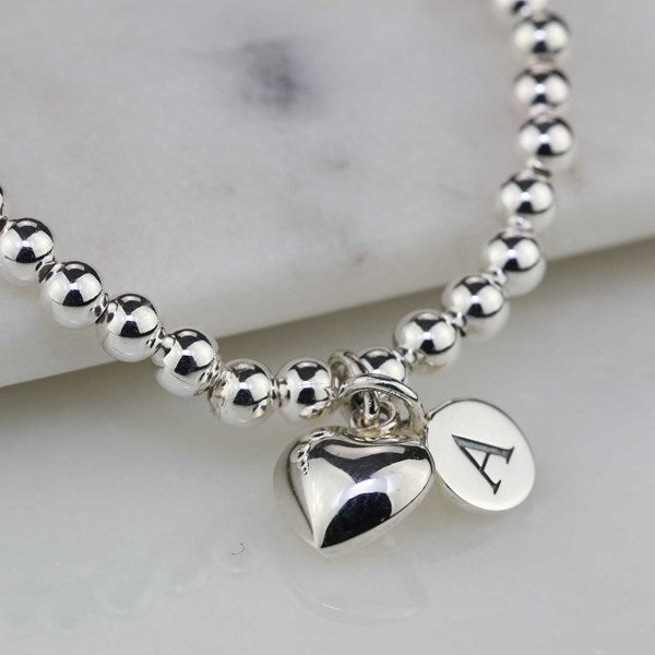 A silver beaded bracelet with a polished heart charm and a round initial pendant engraved with the letter 'A', displayed on a light marble surface.