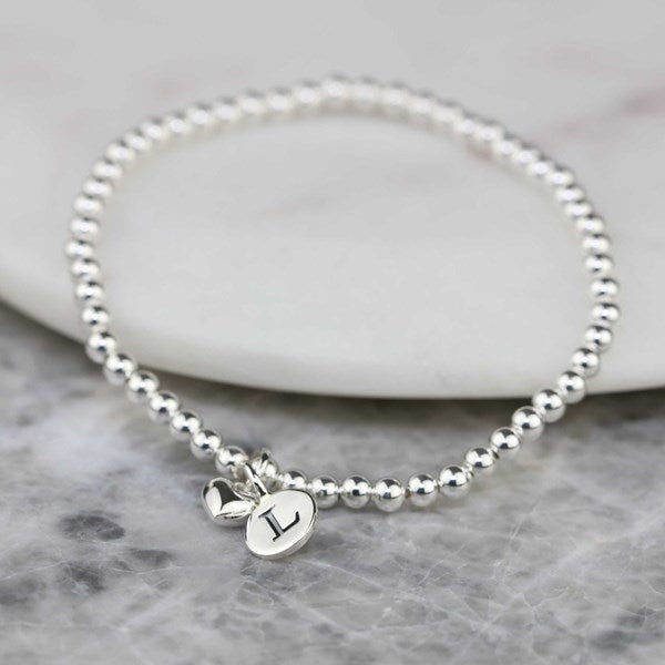 A silver beaded bracelet with two small heart-shaped charms, one engraved with the letter 'L', displayed on a white stone slab over a gray marble surface.