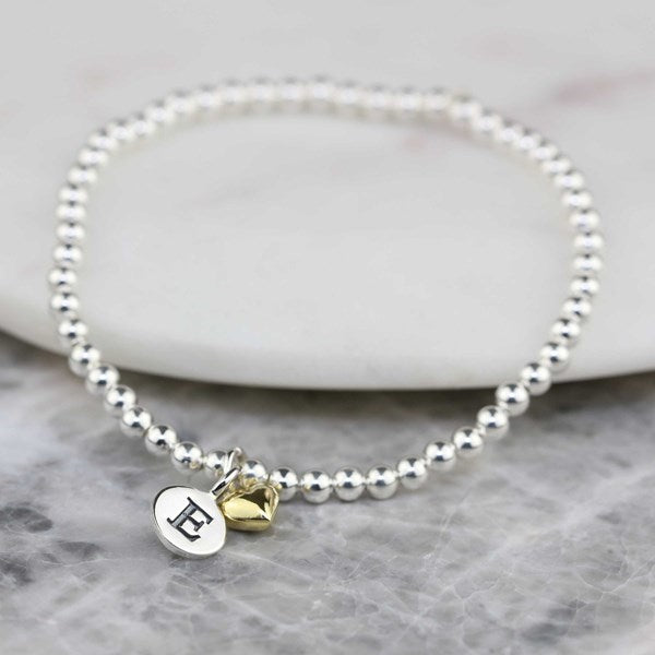 A silver beaded bracelet with a round silver disc charm engraved with the letter 'E' and a small gold heart charm, displayed on a white stone slab over a gray marble surface.