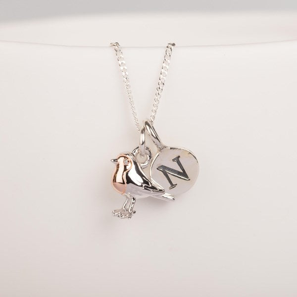 A silver pendant necklace featuring a bird charm with a rose gold accent and a round silver disc engraved with the letter 'N', displayed on a clean white background.