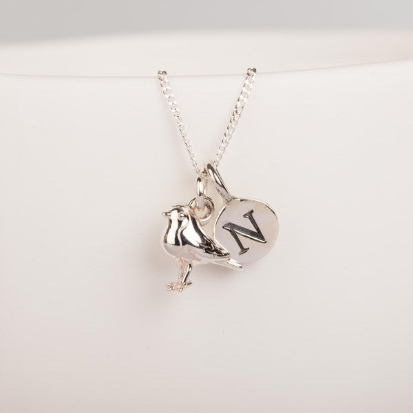 A silver pendant necklace featuring a bird and a heart-shaped charm engraved with the letter 'N', displayed on a clean white background.