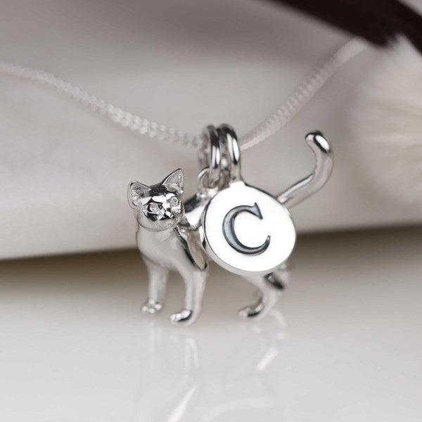 A silver cat pendant necklace with a round initial 'C' charm, shown close-up on a white surface with a blurred white background.