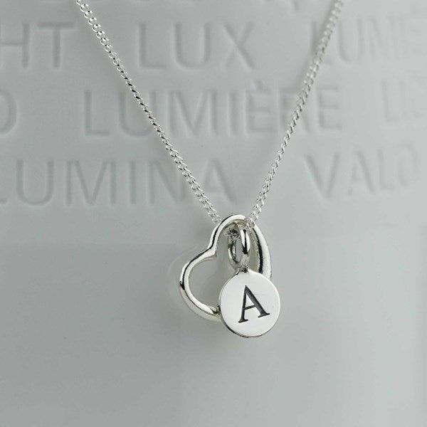 A silver necklace with a heart pendant, and a flat round disc engraved with the letter 'A', displayed against a light gray background with embossed text.