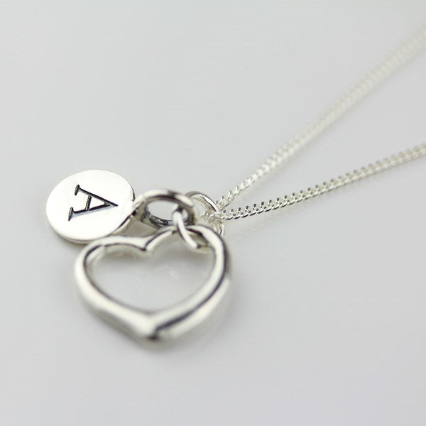A silver necklace with a heart pendant and a round disc charm engraved with the letter 'A', shown on a light gray background.