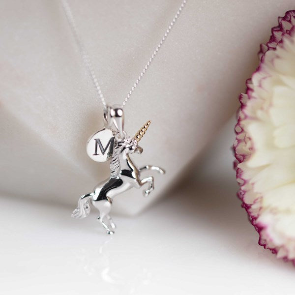 A silver unicorn pendant necklace with a gold horn and a round initial 'M' charm, displayed on a white jewelry stand next to a white flower with purple edges.
