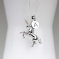 A silver unicorn pendant necklace with a rearing unicorn design, a small round initial 'A' charm, and a gold horn accent, displayed on a white jewelry bust.