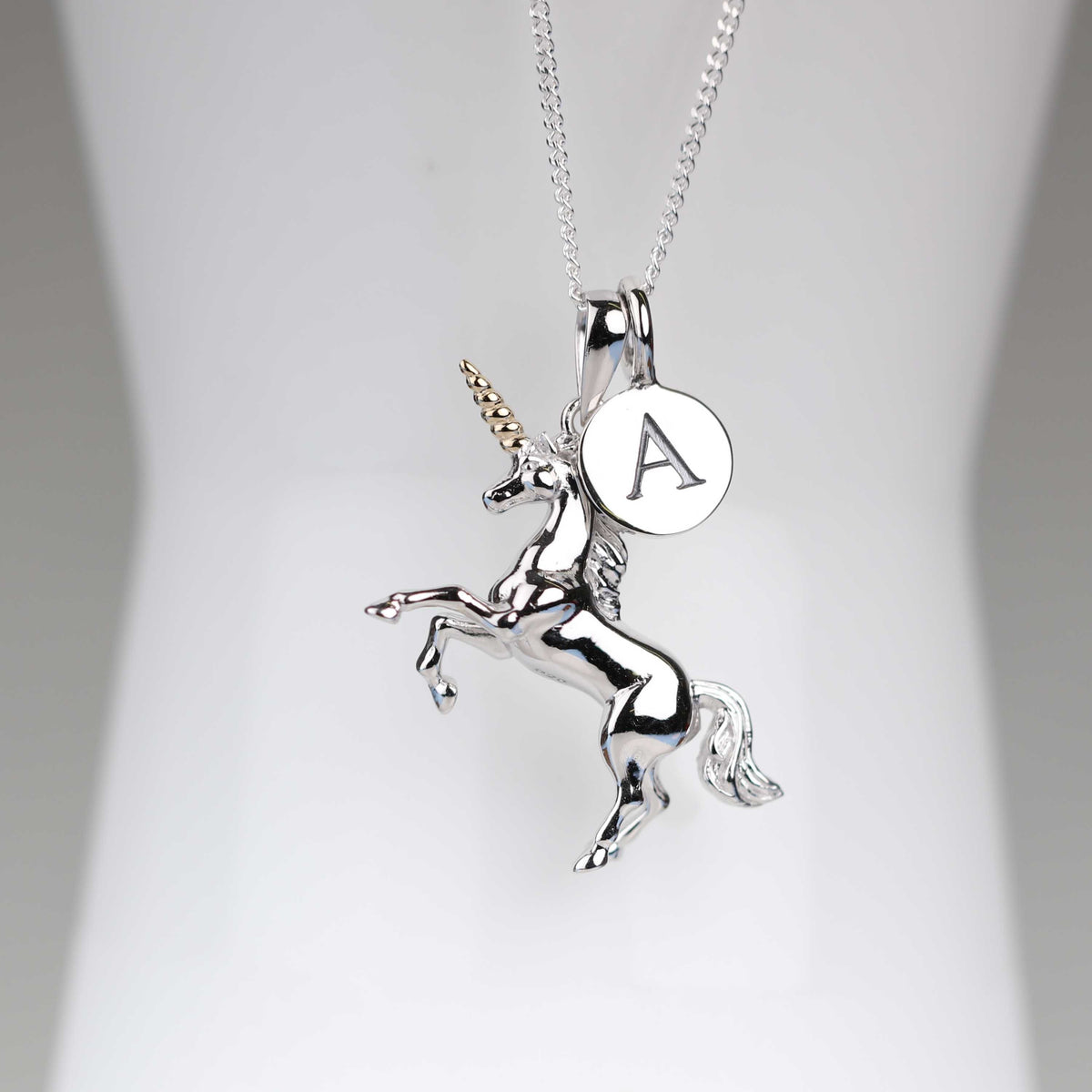 A silver unicorn pendant necklace with a rearing unicorn design, a small round initial 'A' charm, and a gold horn accent, displayed on a white jewelry bust.