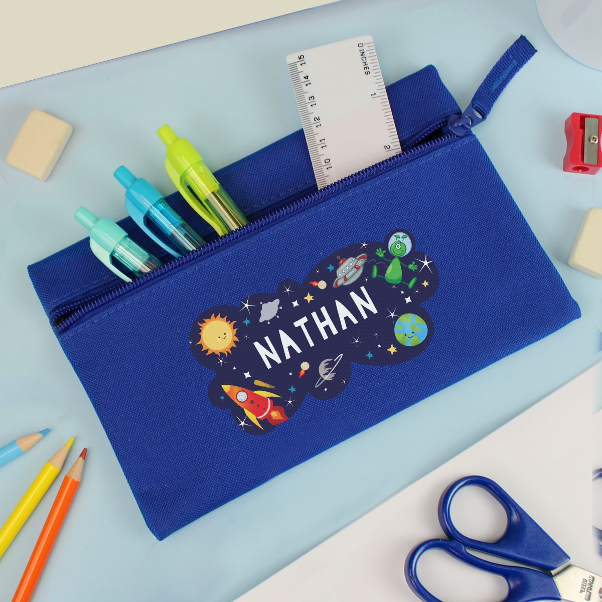 Blue pencil case with the name 'Nathan' and a space theme design on a desk with stationery items