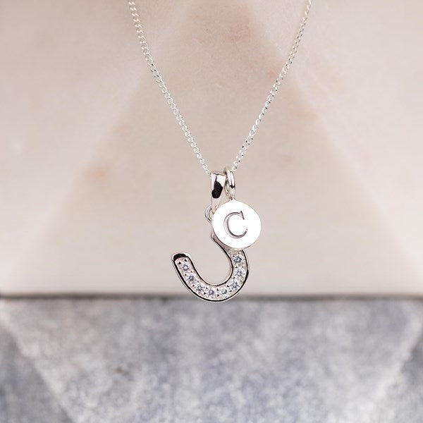 A silver necklace with a horseshoe-shaped pendant set with small clear stones and a round disc charm engraved with the letter 'C', displayed against a soft beige and gray background.