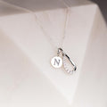 A silver necklace with a delicate chain, featuring a double pendant: a round silver disc engraved with the letter 'N' and a teardrop-shaped pendant with a row of clear crystals along one side, displayed on a white geometric surface.