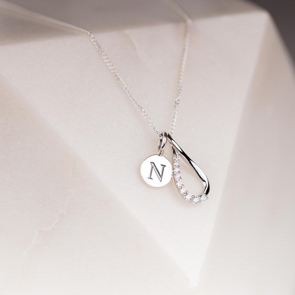 A silver necklace with a delicate chain, featuring a double pendant: a round silver disc engraved with the letter 'N' and a teardrop-shaped pendant with a row of clear crystals along one side, displayed on a white geometric surface.