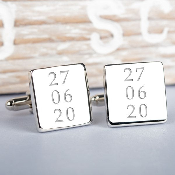 A pair of square silver-toned cufflinks on a light grey surface. Both cufflink faces are engraved with a date.