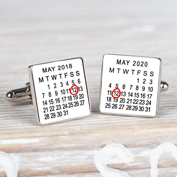 A pair of square silver-toned cufflinks on a light wood surface. Both cufflink faces are white with a printed calendar showing May 2018 and May 2020 with the 12th highlighted in red.