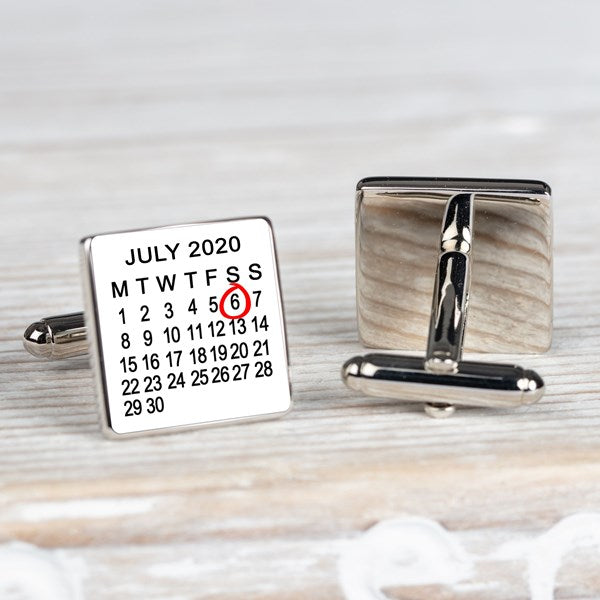 A pair of square silver-toned cufflinks on a light wood surface. One cufflink face is white with a printed calendar showing July 2020 and the 6th highlighted in red.  The other is showing the back of the cufflink.