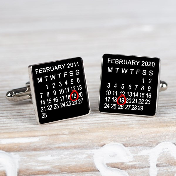 A pair of square silver-toned cufflinks on a light wood surface. Both cufflink faces are black with a printed calendar showing February 2011 and February 2020 and the 19th highlighted in red.