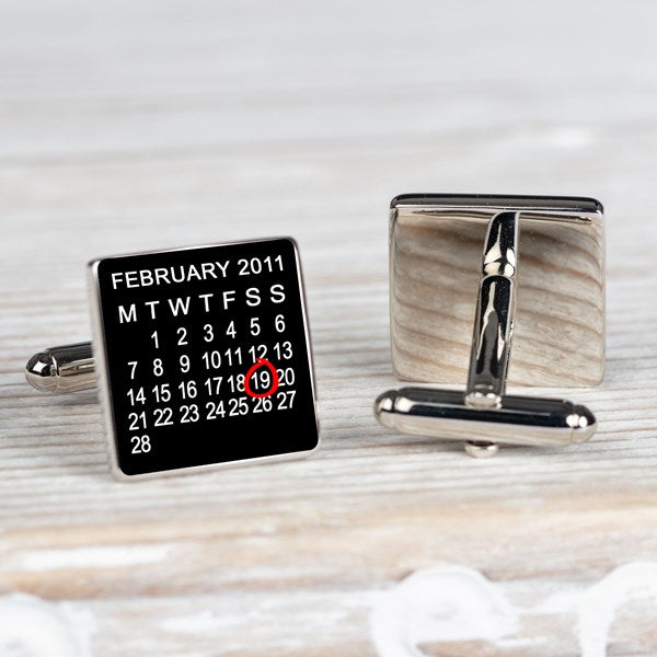 A pair of square silver-toned cufflinks on a light wood surface. One cufflink face is black with a printed calendar showing February 2011 and the 19th highlighted in red. The other is showing the back of the cufflink.