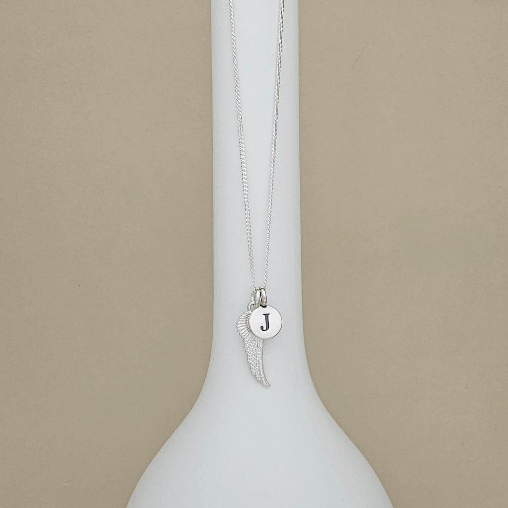 A silver necklace with a wing and round initial pendant displaying the letter 'J', displayed on a white ceramic vase against a beige background.