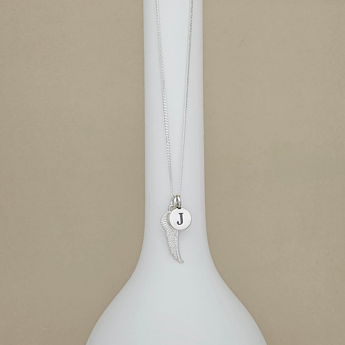A silver necklace with a wing and round initial pendant displaying the letter 'J', displayed on a white ceramic vase against a beige background.