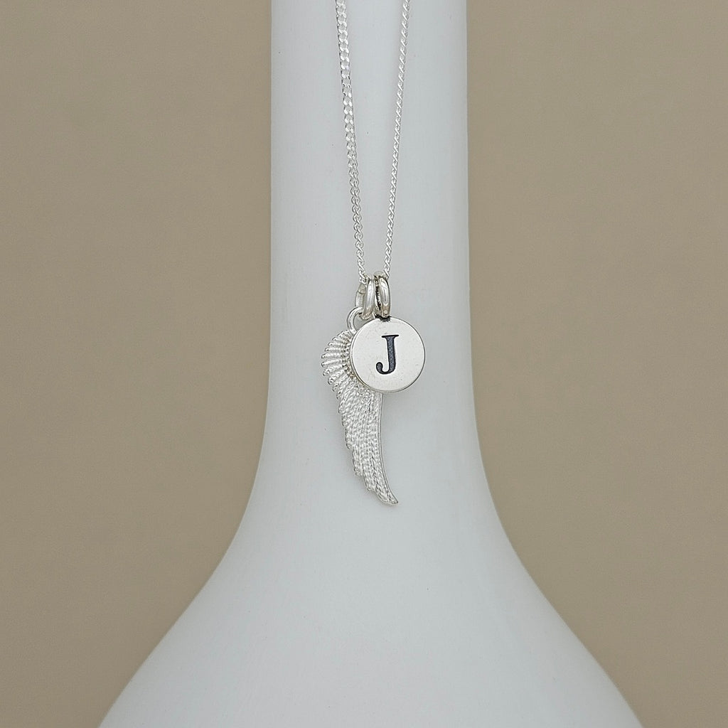 A silver necklace with a wing-shaped pendant and a round initial 'J' disc charm, displayed on a white ceramic vase against a beige background.