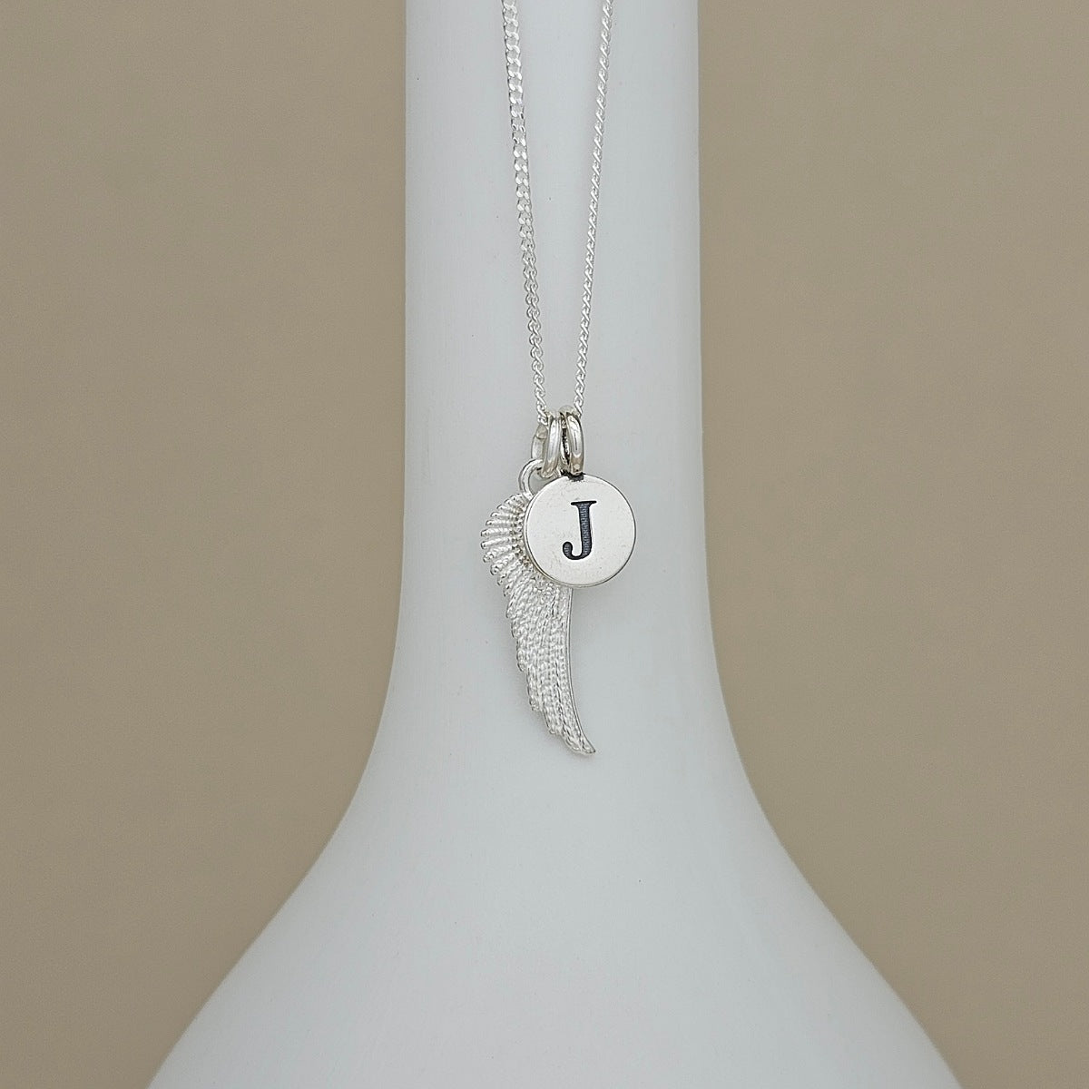 A silver necklace with a wing-shaped pendant and a round initial 'J' disc charm, displayed on a white ceramic vase against a beige background.