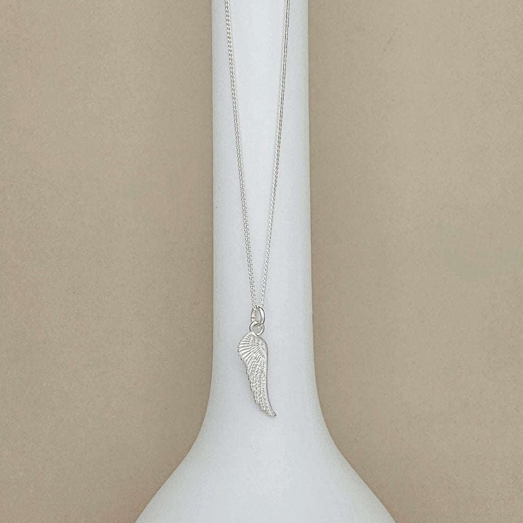 A silver wing pendant necklace displayed on a white neck mannequin against a neutral beige background.