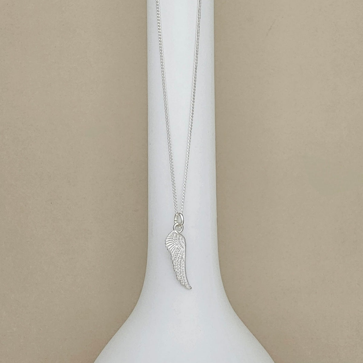 A silver wing pendant necklace displayed on a white neck mannequin against a neutral beige background.