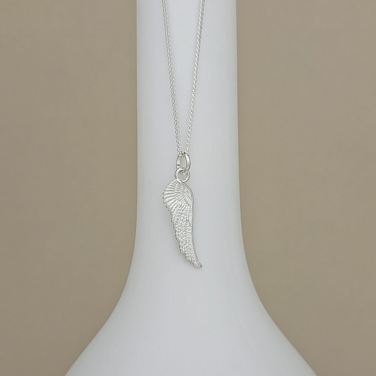 A silver wing pendant necklace displayed on a white neck mannequin against a neutral beige background.