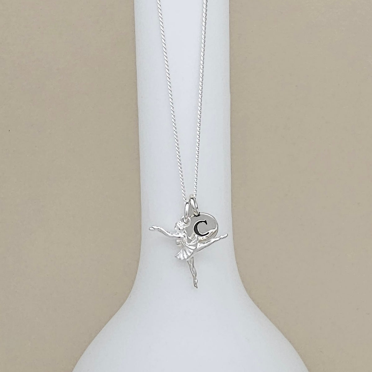 A silver ballerina pendant necklace with a small circular initial 'C' charm, displayed on a white ceramic stand against a neutral beige background.