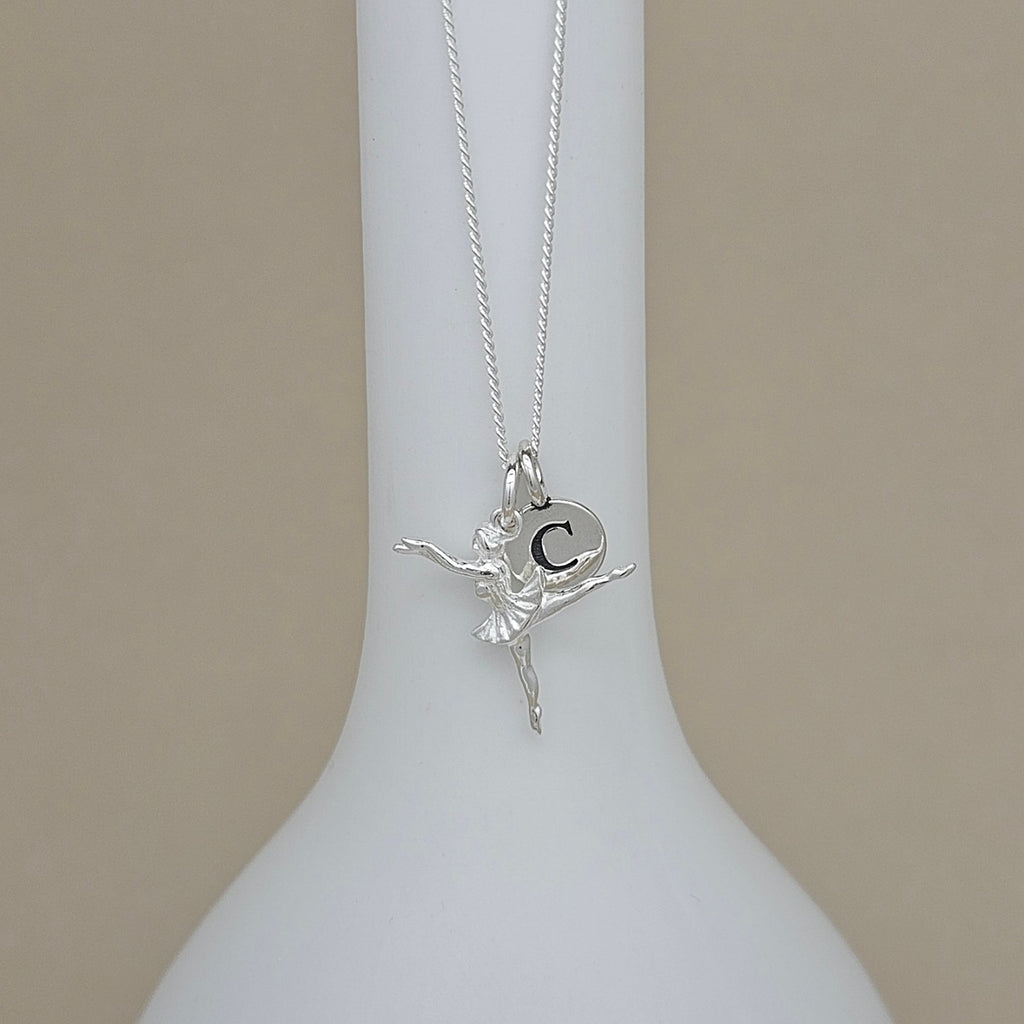 A silver necklace with a ballerina pendant featuring the letter 'C', displayed on a white ceramic stand against a beige background.