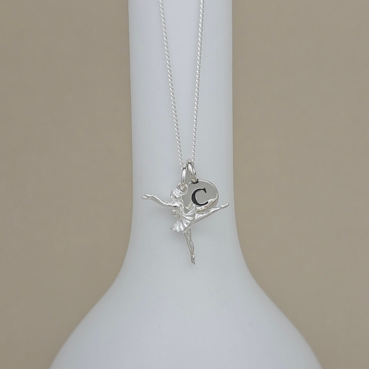 A silver necklace with a ballerina pendant featuring the letter 'C', displayed on a white ceramic stand against a beige background.