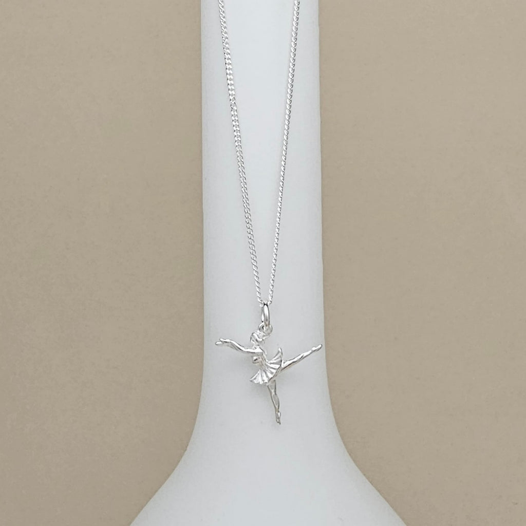 A silver ballerina pendant necklace on a delicate chain, displayed on a white vase against a neutral beige background.