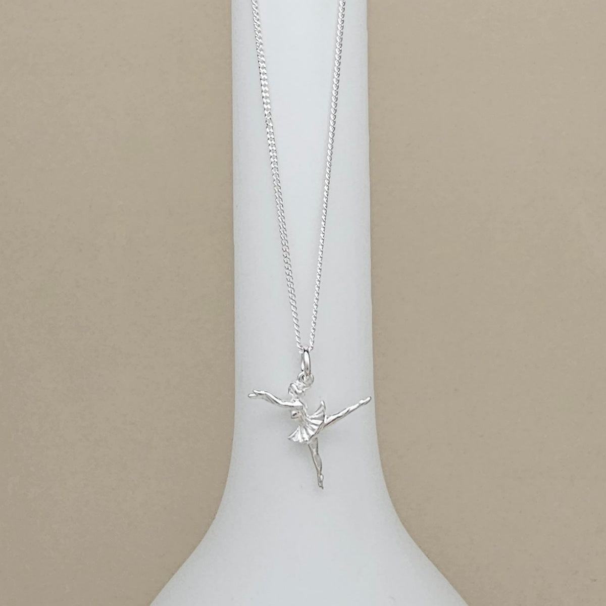 A silver ballerina pendant necklace on a delicate chain, displayed on a white vase against a neutral beige background.