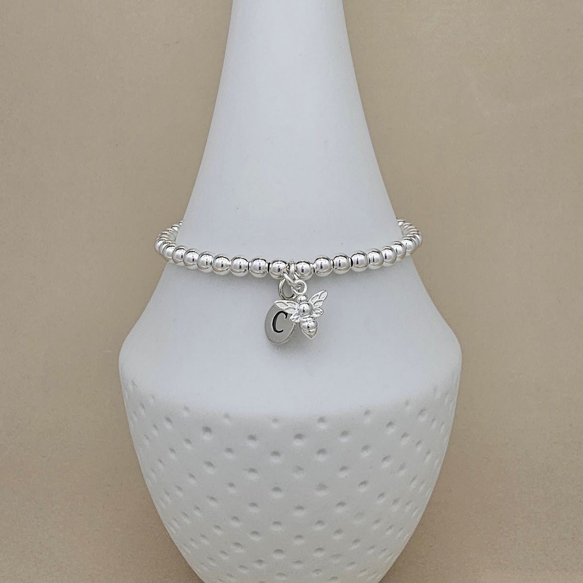A silver beaded bracelet with a small bee charm and a round initial 'C' pendant, displayed on a white ceramic cone stand against a beige background.