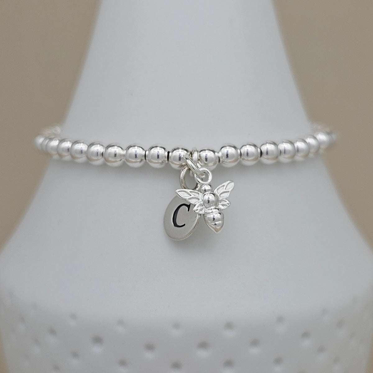 A silver beaded bracelet with a dangling bee charm and a round initial 'C' charm, displayed on a white jewelry bust against a soft beige background.
