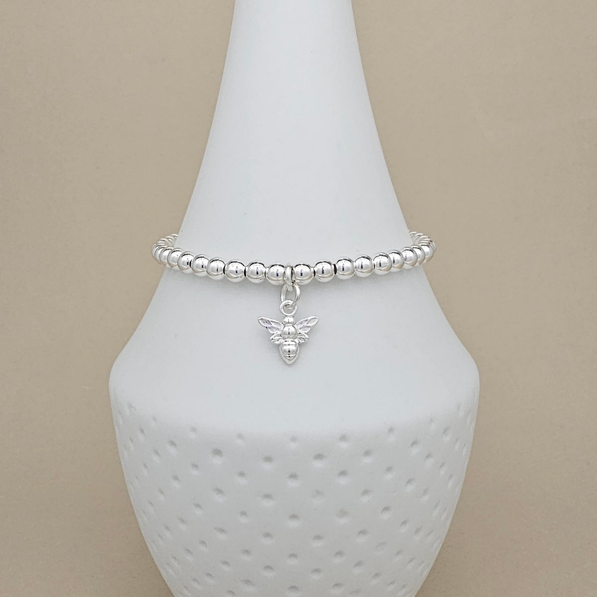 A silver beaded bracelet with a small bee charm, displayed on a white ceramic cone stand against a beige background.