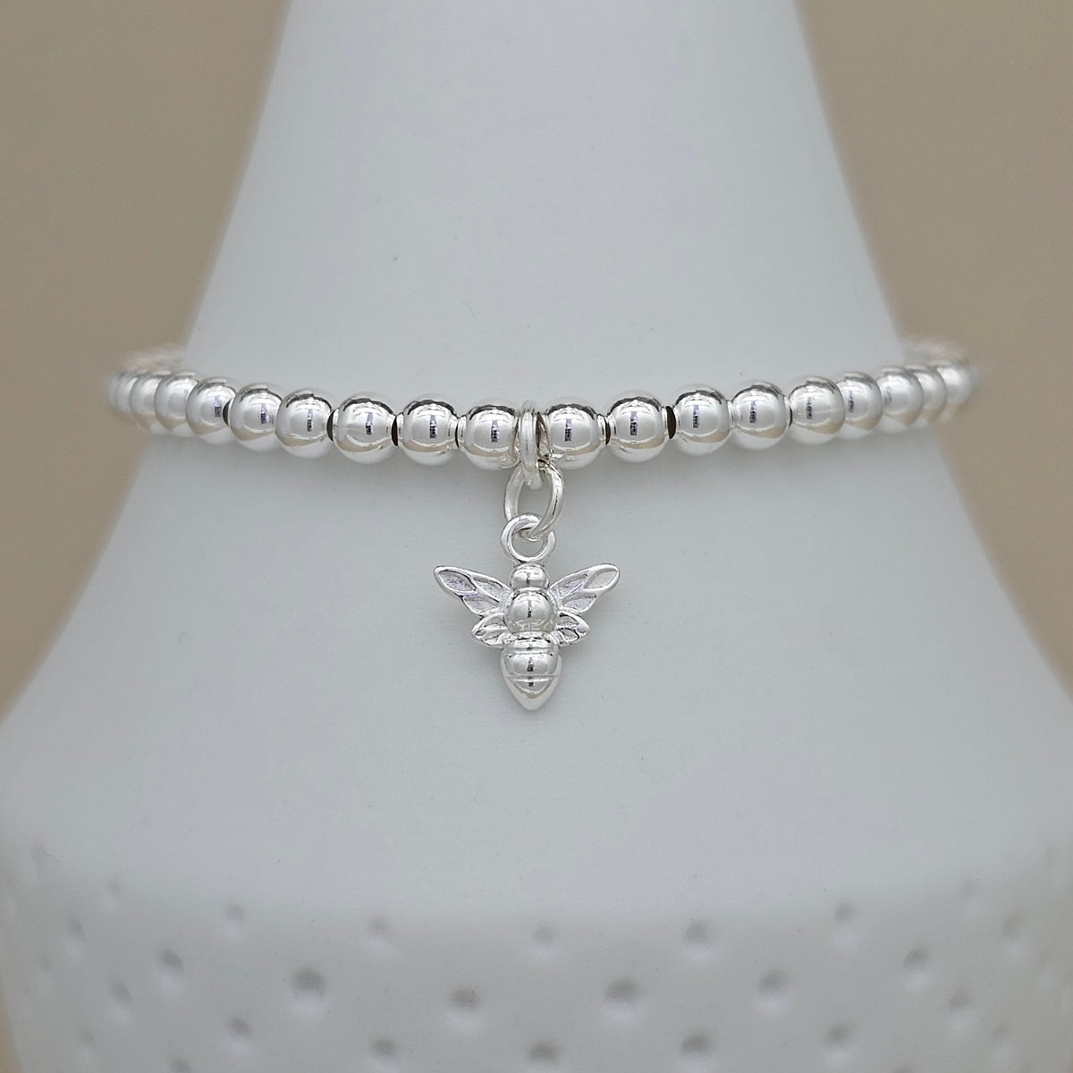 A silver beaded stretch bracelet with a small winged bee charm, displayed on a white cone-shaped jewelry stand against a soft beige background.