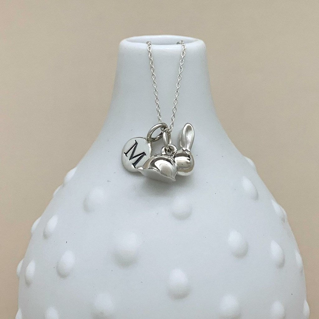 A silver necklace with a delicate chain and three charms, including a heart engraved with the letter 'M', displayed on a white textured vase against a beige background.