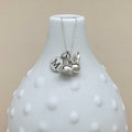 A silver necklace with a delicate chain and three charms, including a heart engraved with the letter 'M', displayed on a white textured vase against a beige background.