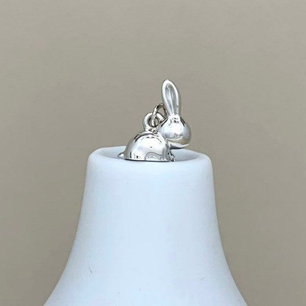 A polished silver bunny charm with a small hanging loop, displayed on top of a white ceramic bell against a solid beige background.