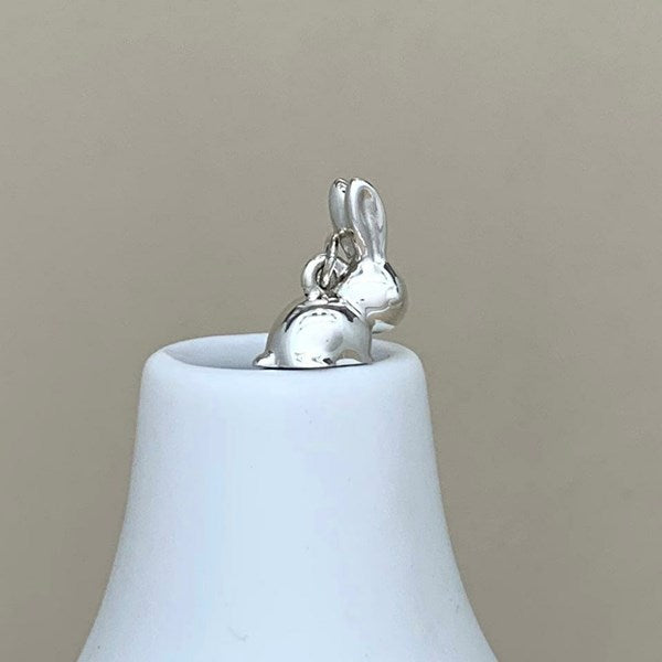 A polished silver bunny charm with long ears, sitting on top of a white bell-shaped ceramic holder against a solid beige background.