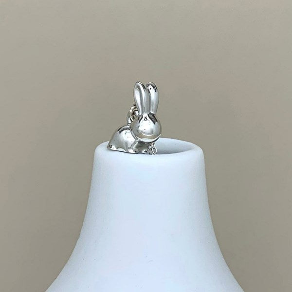 A polished silver bunny charm with long ears, sitting on the rim of a white ceramic bell-shaped container against a solid beige background.