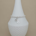 A silver beaded bracelet with a dangling initial 'M' charm and a small angel charm, displayed on a white ceramic vase against a beige background.