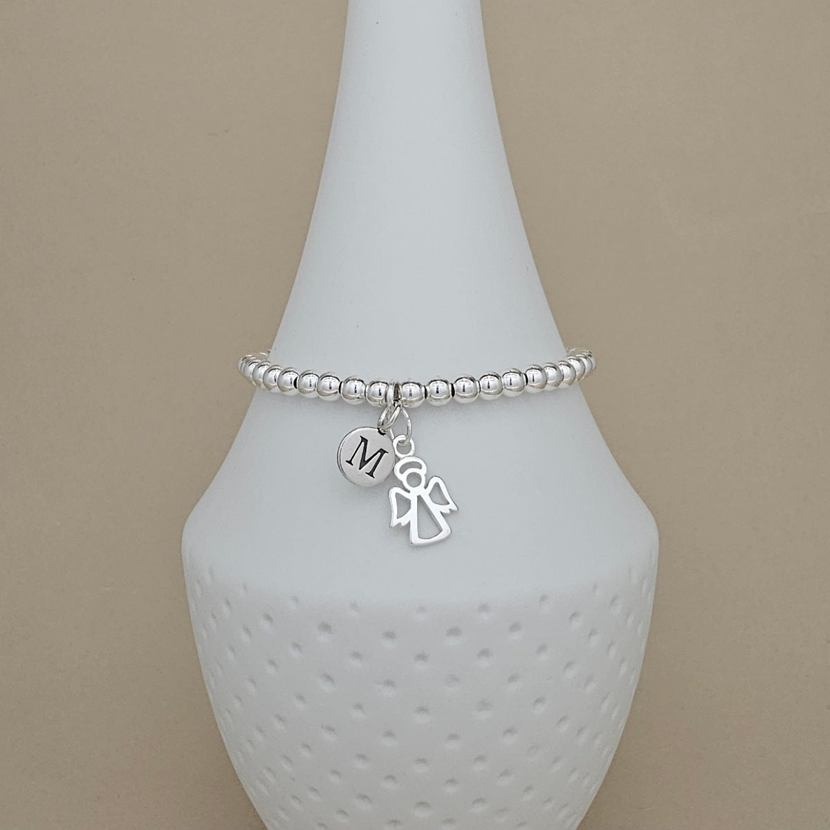 A silver beaded bracelet with a dangling initial 'M' charm and a small angel charm, displayed on a white ceramic vase against a beige background.