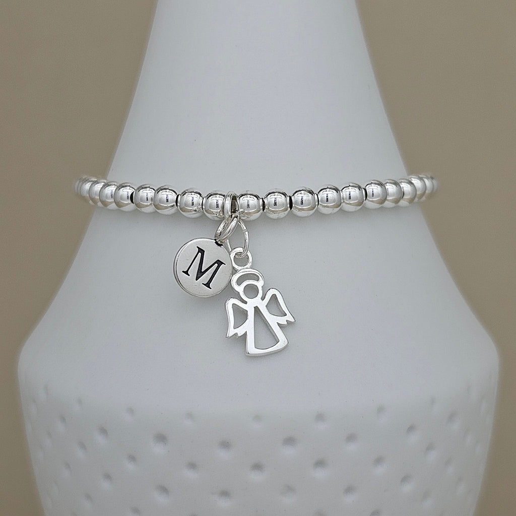 A silver beaded bracelet with a circular initial 'M' charm and an angel silhouette charm, displayed on a white textured cone stand.