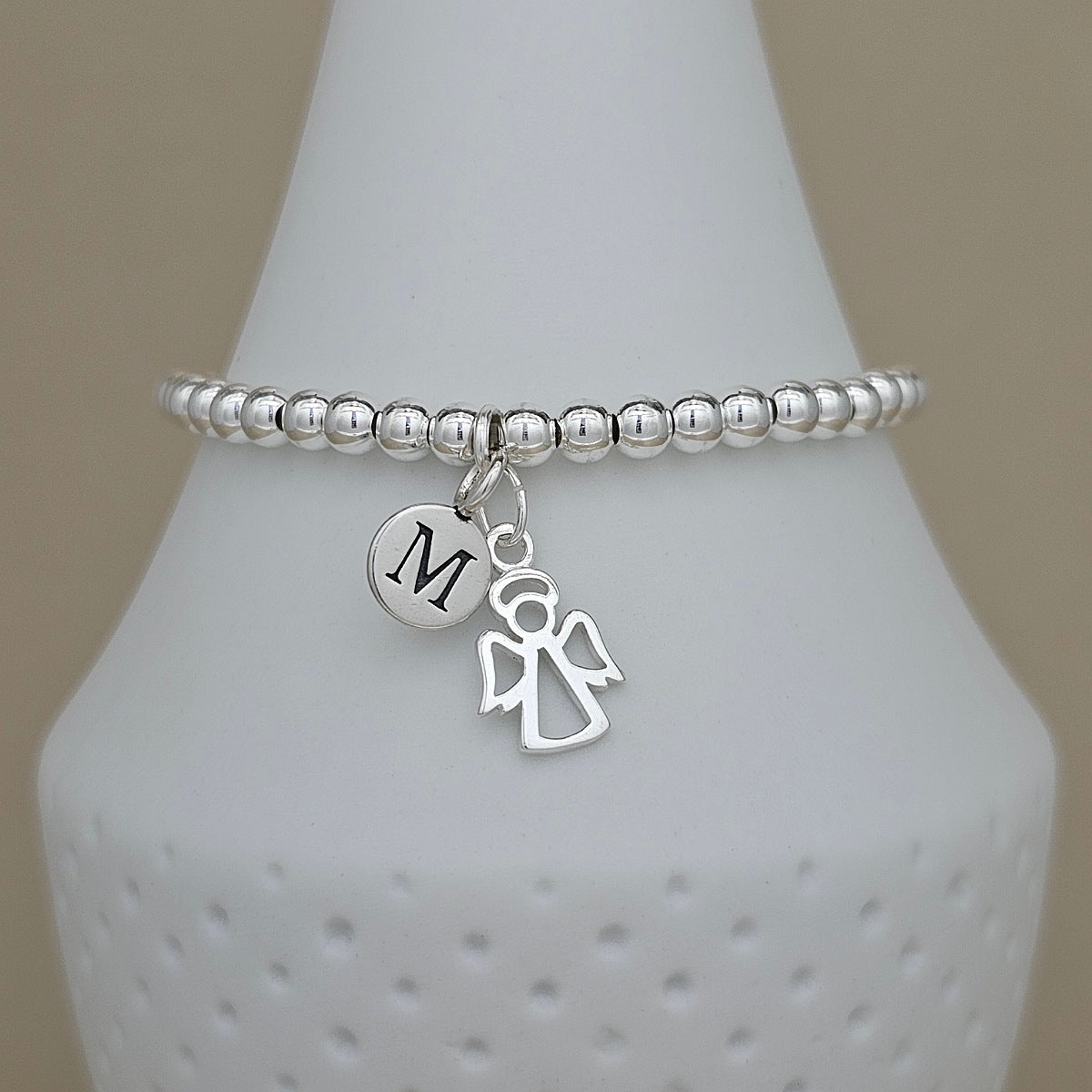 A silver beaded bracelet with a circular initial 'M' charm and an angel silhouette charm, displayed on a white textured cone stand.